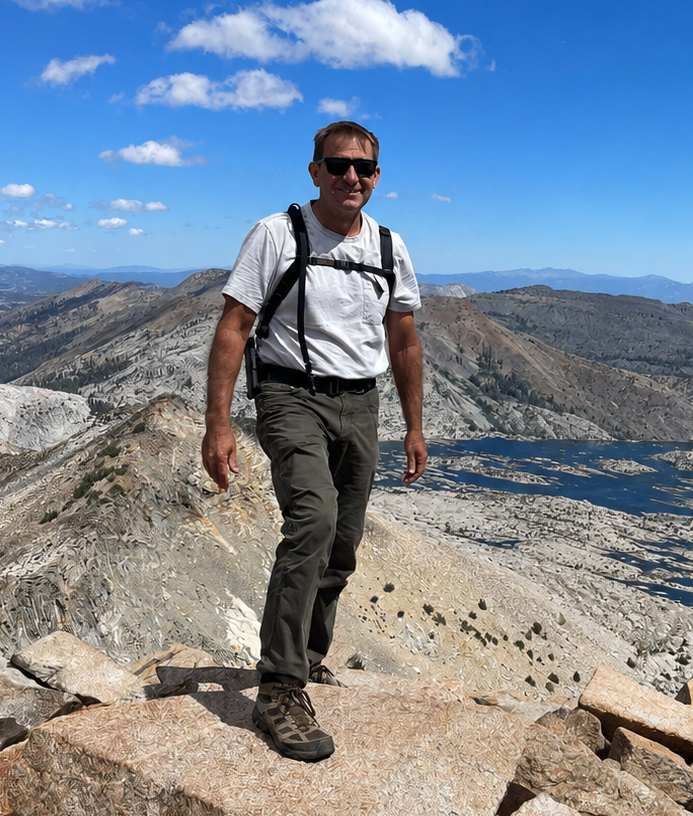 Man hiking on rocky summit with backpack, rugged mountain peaks and bright blue sky
