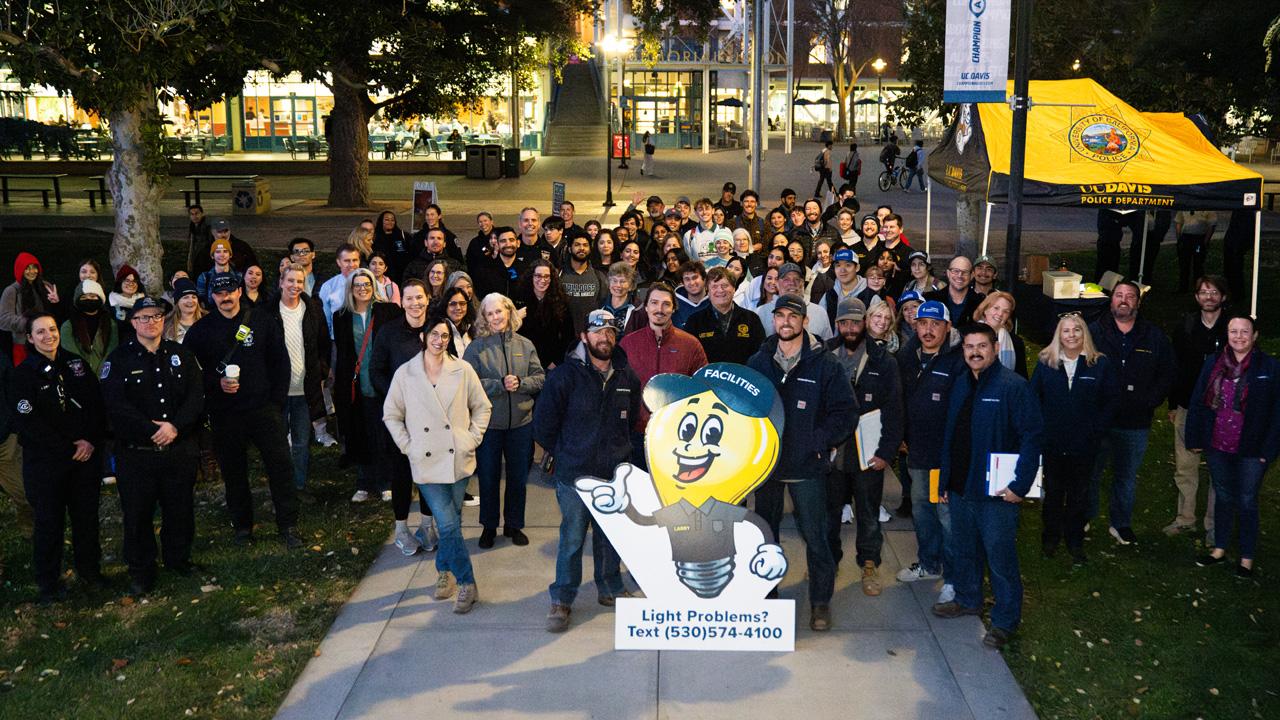 Group photo of UC Davis staff and students outside of the Memorial Union after dark with a cut-out of the Campus Safety Lighting Walk mascot Bulby in front.