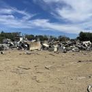 A large pile of broken furniture, metal, and construction debris scattered across a dirt lot, with two trucks parked in the background under a blue sky with scattered clouds.
