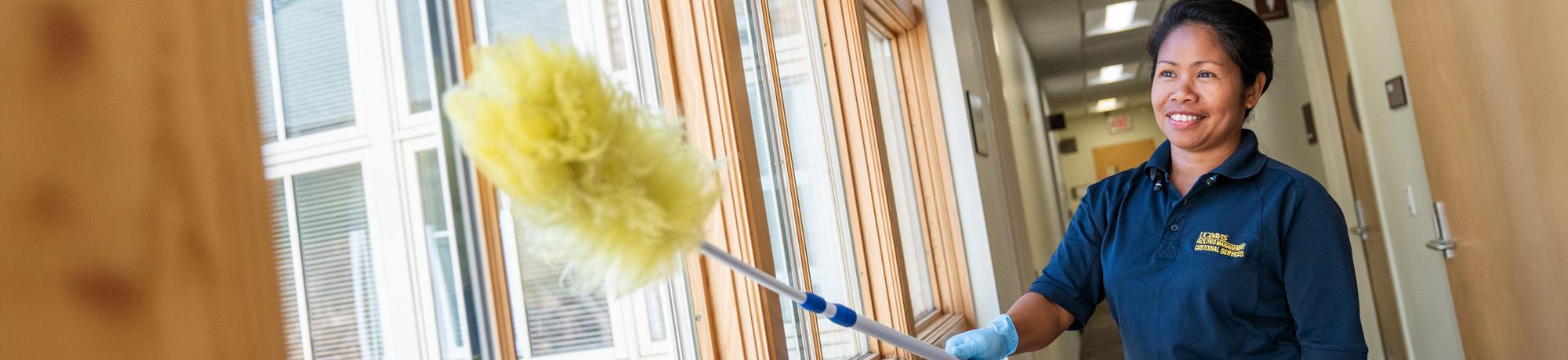 Image of a UC Davis custodian dusting a window sill while smiling and wearing a uniform.